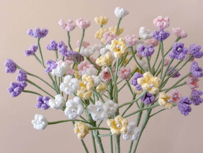 May include: A bouquet of crocheted flowers in shades of purple, pink, white, and yellow. The flowers are arranged in a vase and are a beautiful addition to any home.