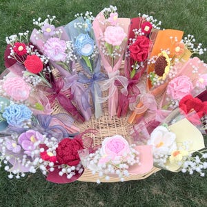 May include: A collection of colorful, crocheted flower bouquets in a woven basket. The bouquets feature various flower types in shades of pink, blue, red, and yellow, wrapped in decorative paper and tied with ribbons. White filler flowers add detail.