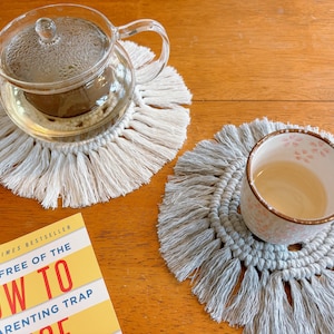 May include: Two crocheted coasters with fringe, one in white and one in gray, with a glass teapot and a ceramic cup on top of them. The teapot and cup are both filled with a light-colored liquid.