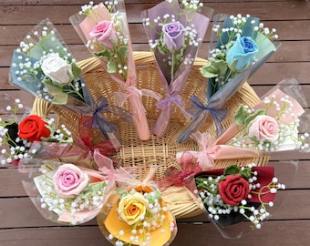 May include: A collection of nine crocheted rose bouquets in various colors, including pink, white, blue, red, and yellow. Each bouquet is wrapped in clear cellophane and tied with a ribbon. The bouquets are arranged in a wicker basket.