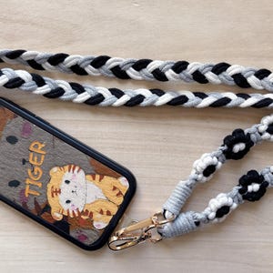 May include: A black and white braided phone strap with black and white flower accents. The strap is attached to a black phone case with a cartoon tiger design and the word "TIGER" printed on it.