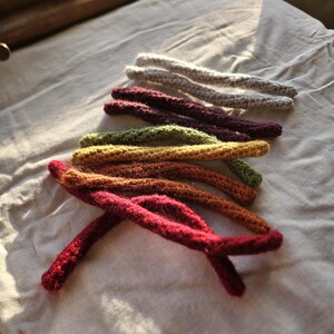 May include: A set of crocheted yarn strands in various colors, including red, orange, yellow, green, purple, and gray. The strands are arranged in a rainbow pattern on a white fabric surface.