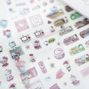 May include: A sheet of pink and white Hello Kitty stickers featuring various images of Hello Kitty, including Hello Kitty with glasses, Hello Kitty with a phone, Hello Kitty with a camera, and Hello Kitty with a star. The stickers are on a clear background.