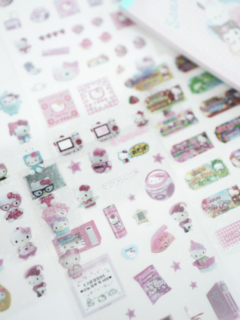 May include: A sheet of pink and white Hello Kitty stickers featuring various images of Hello Kitty, including Hello Kitty with glasses, Hello Kitty with a phone, Hello Kitty with a camera, and Hello Kitty with a star. The stickers are on a clear background.