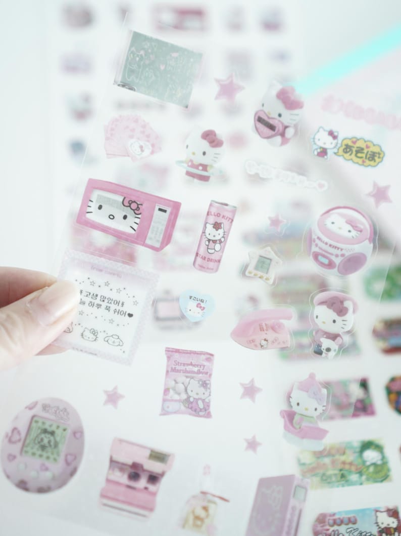 May include: A sheet of clear stickers featuring Hello Kitty characters and objects. The stickers are pink and white and include a microwave, a can of Hello Kitty soda, a boombox, a phone, a star, and a Hello Kitty character holding a bag of marshmallows. The text on the stickers includes "Hello Kitty", "Strawberry Marshmallow", and "오늘은" in Korean.