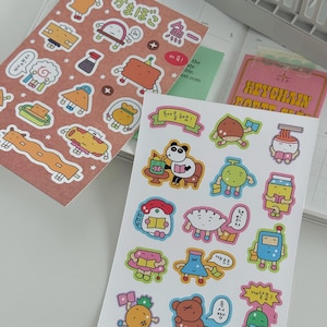 Op de afbeelding: Twee stickervellen met kleurrijke illustraties. Eén vel is bruin met Japanse tekst en stickers met een voedselthema. De andere is wit met stripfiguren die boeken lezen en Koreaanse tekst. Een roze sleutelhanger is ook zichtbaar.