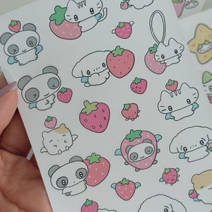May include: Sheet of stickers featuring cute cartoon animals and strawberries. The stickers include pandas, cats, bunnies, and other characters, each with a strawberry or wearing a strawberry costume. The stickers are in various sizes and colors, with a white background.