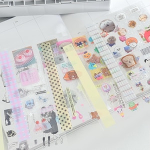 May include: A collection of clear sticker sheets featuring various cartoon characters, food items, and decorative patterns. The stickers are arranged on a white surface, with a light yellow tape and a white storage bin in the background.
