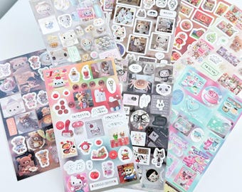 Peut inclure: Plusieurs planches d'autocollants colorés avec des personnages de dessins animés et des motifs variés, notamment des ours, des chats et des illustrations d'aliments. Les autocollants sont disposés sur une surface blanche.