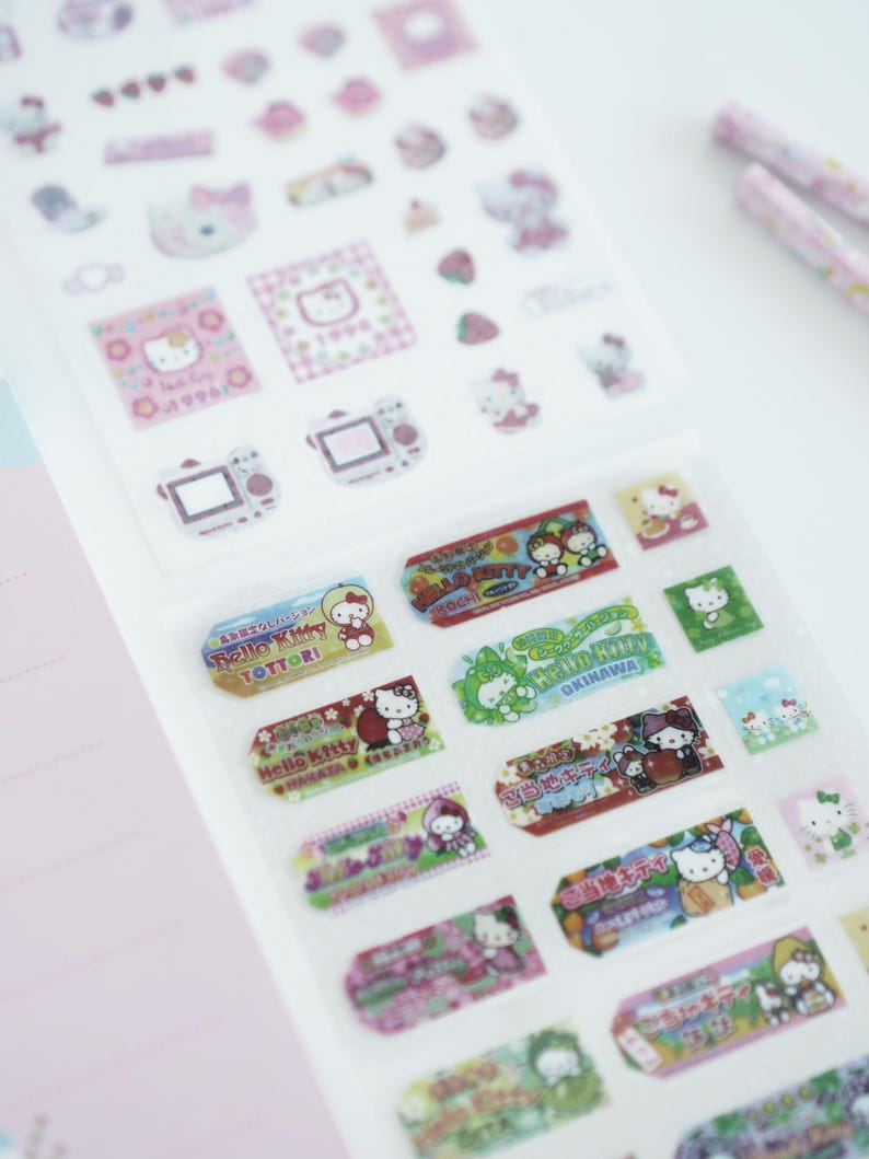 May include: A sheet of Hello Kitty stickers featuring various designs, including Hello Kitty in different outfits and locations, with text in Japanese. The stickers are on a white background.