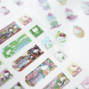 May include: A sheet of colorful Hello Kitty stickers featuring various designs, including Hello Kitty with a pineapple, Hello Kitty with a cherry, and Hello Kitty with a green leaf. The stickers are on a white background.
