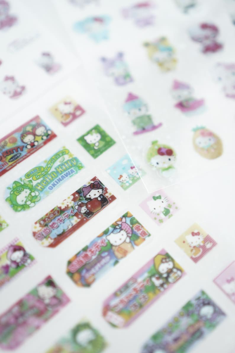 May include: A sheet of colorful Hello Kitty stickers featuring various designs, including Hello Kitty with a pineapple, Hello Kitty with a cherry, and Hello Kitty with a green leaf. The stickers are on a white background.