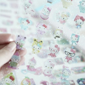 May include: A sheet of clear stickers featuring Hello Kitty characters in various poses and outfits. The stickers are on a white background with a glittery effect.