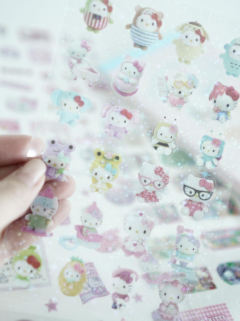 May include: A sheet of clear stickers featuring Hello Kitty characters in various poses and outfits. The stickers are on a white background with a glittery effect.