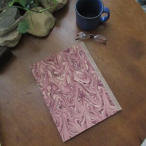 May include: A book with a marbled cover in shades of pink and cream. The cover has a swirling pattern. The book is lying on a wooden table.