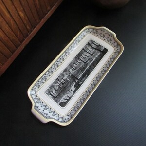 May include: Rectangular serving tray with a black and white landscape design in the center. The tray has a cream-colored border with a floral pattern and gold trim. The tray has handles on each end.