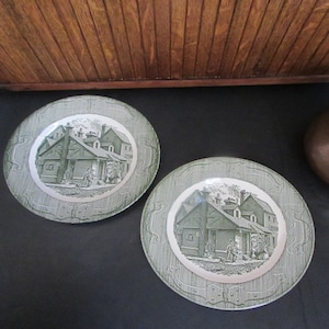 May include: Two green and white ceramic plates with a vintage design featuring a black and white illustration of a building with a chimney and people walking in front of it.