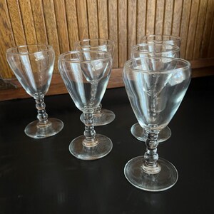 May include: Six clear glass stemmed drinking glasses with a beaded stem. The glasses are arranged in a row on a black surface.