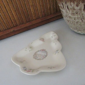 May include: A white ceramic Christmas tree-shaped dish with a floral design and a small, circular design in the center. The dish is decorated with pink, blue, and green flowers.