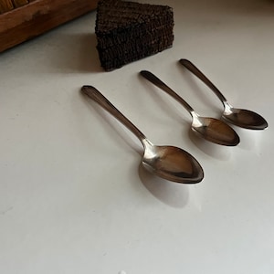 May include: Three silver spoons with oval-shaped bowls, arranged in a row on a white surface.