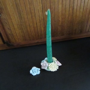 May include: A porcelain candle holder with a green candle and floral accents. The holder features a ring of pastel-colored flowers, including pink, yellow, and blue.