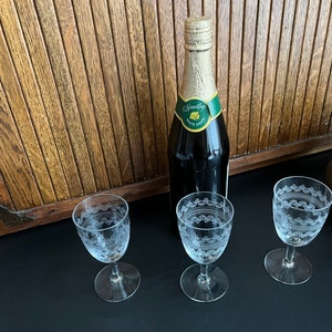 May include: Three clear glass stemmed drinking glasses with etched designs and a bottle of sparkling white wine on a black surface.