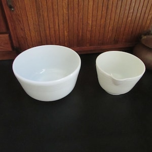 May include: Two white mixing bowls, one larger than the other, on a black surface. The larger bowl is round and has a slight lip around the top. The smaller bowl is also round and has a pouring spout on one side.