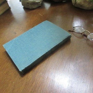May include: A blue hardcover book with a worn cover, lying on a wooden surface. A pair of eyeglasses is lying on the surface to the right of the book.