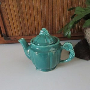 May include: A small teal ceramic teapot with a floral design on the lid and handle. The teapot has a rounded body and a spout on one side.