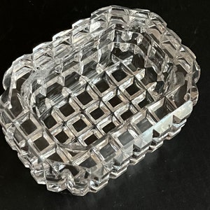 May include: A clear glass ashtray with a square shape and a grid pattern. The ashtray is made of cut glass and has a raised edge.