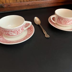 May include: Two white teacups and saucers with a red and white scenic design, along with a silver spoon. The cups and saucers are placed on a dark surface, suggesting a tea set.