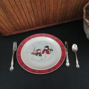 May include: A white ceramic dinner plate with a red and white border featuring a winter scene with two snowmen. The plate is set on a black tablecloth with a silver fork, knife, and spoon.