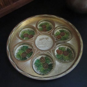 May include: A gold metal tray with six round indentations, each featuring a painted design of pine cones and branches. The tray has a gold rim and a white inscription that reads "The Smokies North Carolina".