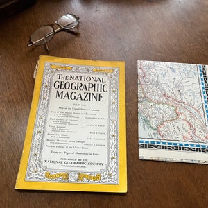 May include: A yellow National Geographic Magazine from July 1956 with the text "The National Geographic Magazine" and "Published by the National Geographic Society" on the cover. The magazine is lying on a wooden table next to a folded map.