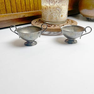 May include: Two small, antique silver-toned sauce bowls with handles. The bowls have a hammered texture and sit on a small pedestal base. The handles are curved. The bowls are displayed on a white surface.