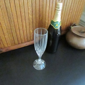 May include: A clear glass champagne flute with a fluted design sits on a black surface next to a bottle of sparkling wine with a green label. The bottle is labeled "Welschriesling"