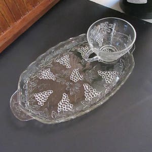 May include: A clear glass serving tray with a grapevine design. The tray has two handles and a matching cup.