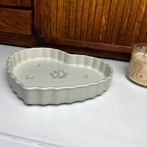 May include: A light green, heart-shaped ceramic dish with a scalloped edge and floral pattern. A glass candle holder with an orange and white design is next to the dish.