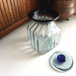 May include: A clear glass jar with a blue rim and a matching lid. The jar has a ribbed design and sits on a white surface. The lid is a small, round disc with a blue knob.