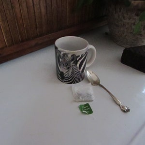 May include: A white ceramic mug with a black and white zebra print design. The mug is filled with a light brown liquid. A silver spoon and a green tea bag with the text "Pure Green Tea" are on a white surface.