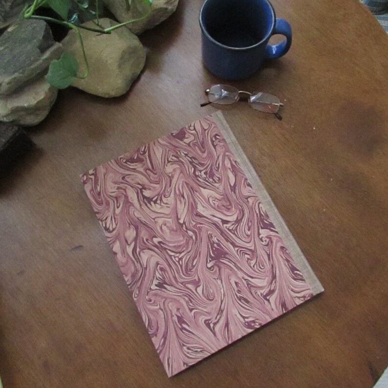 May include: A book with a marbled cover in shades of pink and cream. The cover has a swirling pattern. The book is lying on a wooden table.