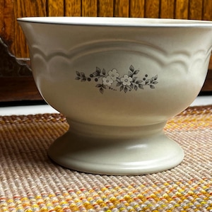 May include: Cream-colored pedestal bowl with a floral design. The bowl has a scalloped rim and a decorative floral pattern. The bowl is made of ceramic and is suitable for serving or display.