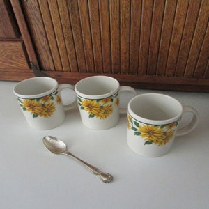 May include: Three white ceramic mugs with a yellow sunflower pattern and green leaves. The mugs have a handle on the right side. A silver teaspoon is in the foreground.