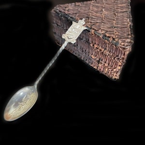 May include: A silver spoon with an ornate handle featuring a crest and a crown. The spoon is lying on a brown woven surface.