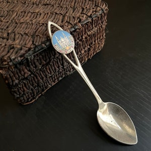 May include: A silver souvenir spoon with a blue and white enamel emblem featuring a church and the text "MARTYRS SHRINE". The spoon is next to a dark brown textured object.