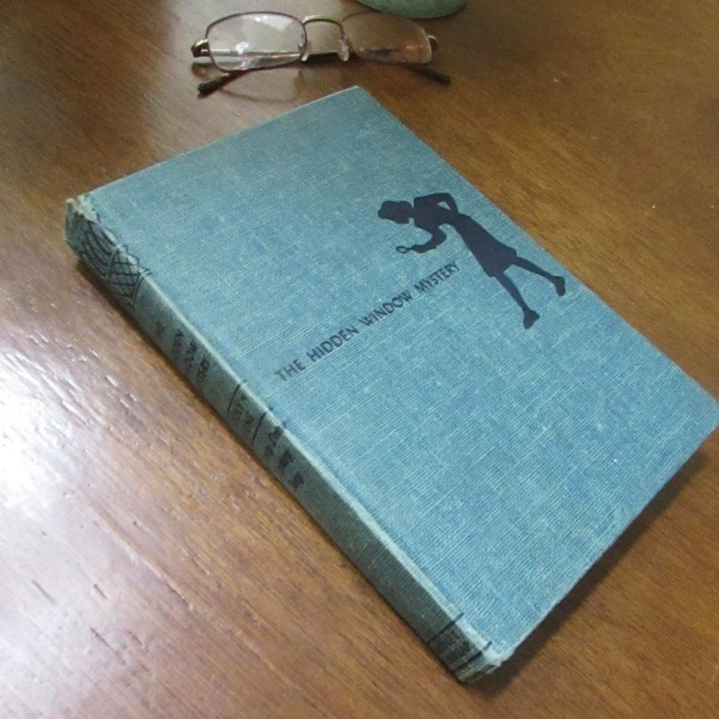 May include: A blue hardcover book with the title "The Hidden Window Mystery" printed on the cover. A silhouette of a person holding a magnifying glass is featured on the cover.