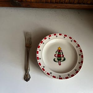 May include: A white ceramic plate with a red and green Christmas tree design. The plate has a red and white border with a repeating pattern of 'S's. A silver fork is placed to the left of the plate.