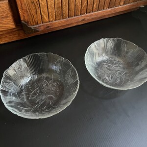 May include: Two clear glass bowls with a leaf design. The bowls have a scalloped edge and are made of clear glass. The leaf design is etched into the glass. The bowls are on a black surface.