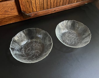May include: Two clear glass bowls with a leaf design. The bowls have a scalloped edge and are made of clear glass. The leaf design is etched into the glass. The bowls are on a black surface.