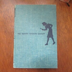 May include: A blue book with the title "The Hidden Window Mystery" printed on the cover. A silhouette of a person holding a magnifying glass is shown on the cover.